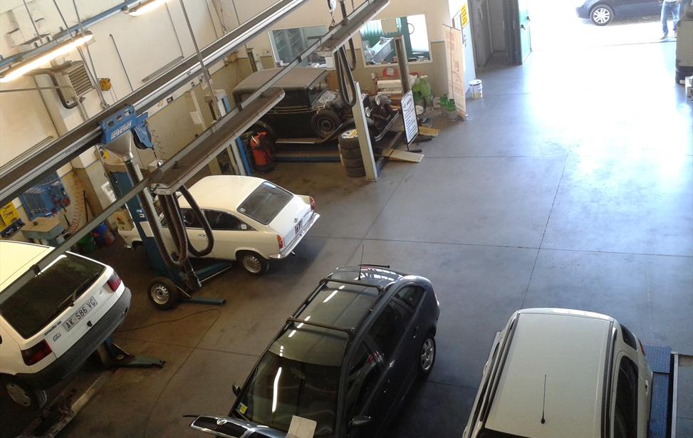Officina vista dall'alto con auto su ponti sollevatori, una coupé bianca d'epoca e una vettura anni Trenta sul fondo
