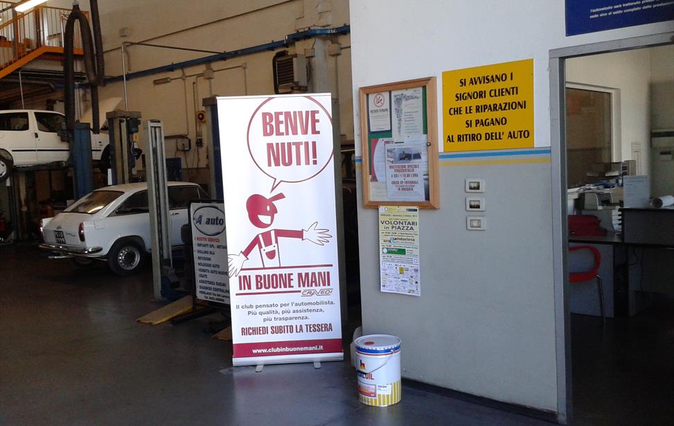 Officina con auto bianca d'epoca, ponti sollevatori, banner «Benvenuti! In buone mani» e cartello giallo sui pagamenti