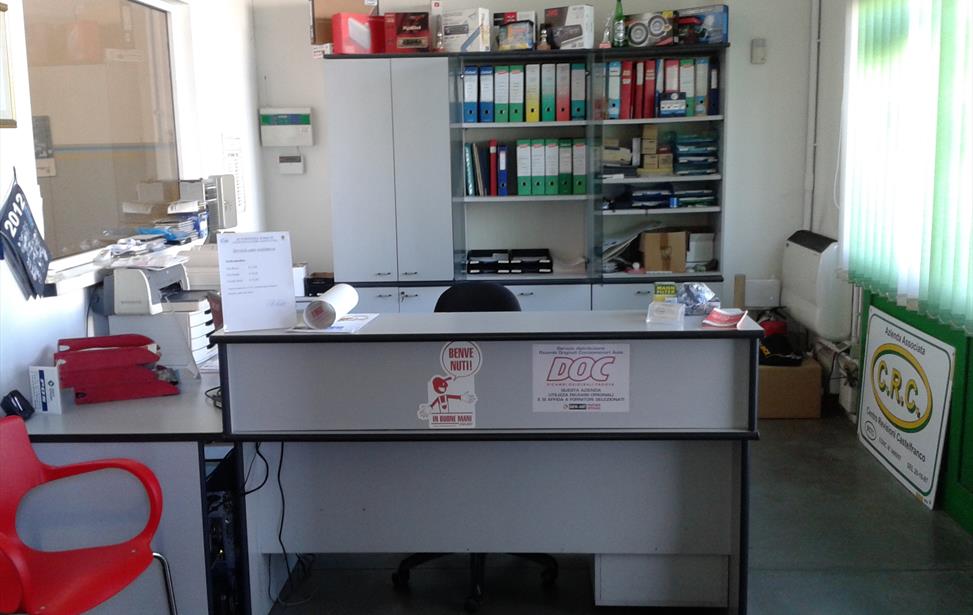 Bancone d'accettazione con adesivi «Benvenuti!» e «DOC», scaffali di raccoglitori colorati e cartello «C.R.C.» in ufficio